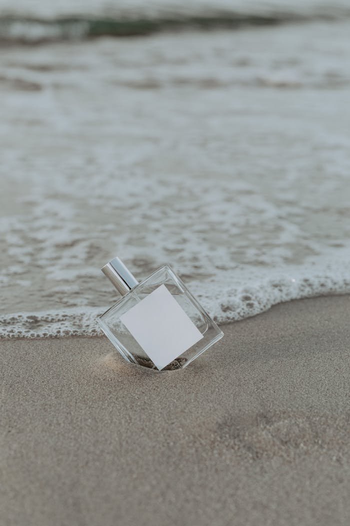 A minimalist perfume bottle resting on sand by the ocean, with gentle waves approaching.